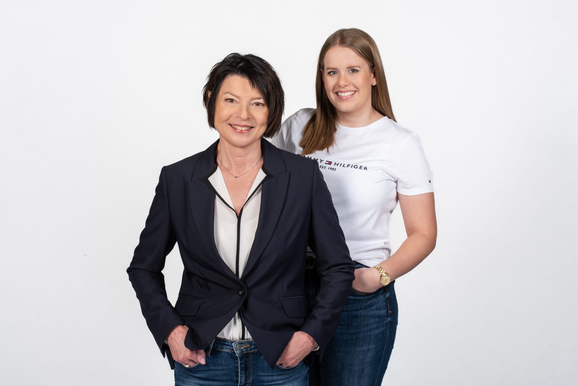 Vor einem weißen Hintergrund stehen zwei Frauen. Die eine trägt einen schwarzen Blazer über einer hellen Bluse, die andere ein weißes T-Shirt von Tommy Hilfiger.